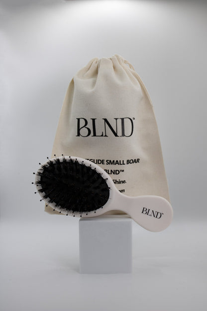 White hairbrush with black boar bristles and a beige drawstring bag labelled 'BLND' on a white background.